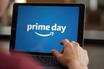 A hand touching a tablet screen displaying the Amazon Prime Day logo on a blue background.