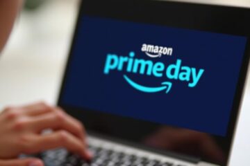 A hand touching a tablet screen displaying the Amazon Prime Day logo on a blue background.