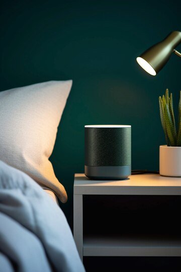 A nightstand with a smart speaker, desk lamp, and small plant next to a bed with a white pillow against a dark green wall.