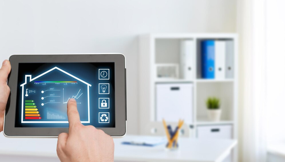 Hand holding tablet displaying smart home energy monitoring interface with house icon, temperature controls, and energy efficiency graph on a minimalist white office background